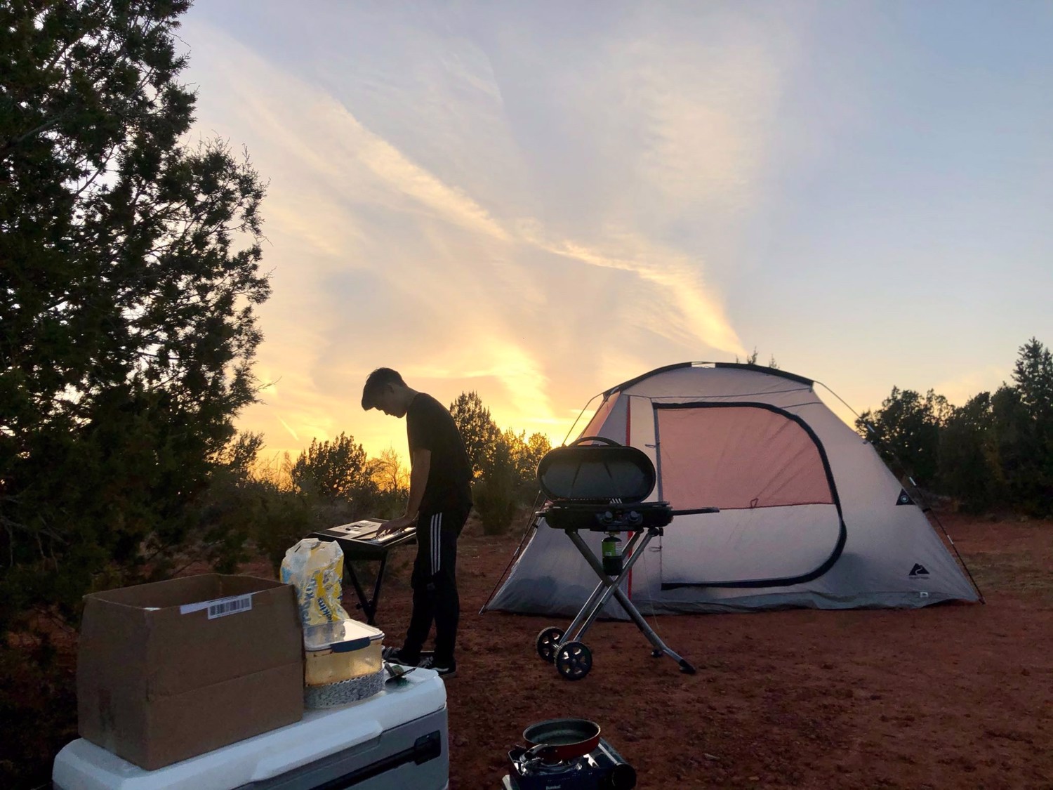 A camper prepares food beside a tent as the sky glows near sunset.