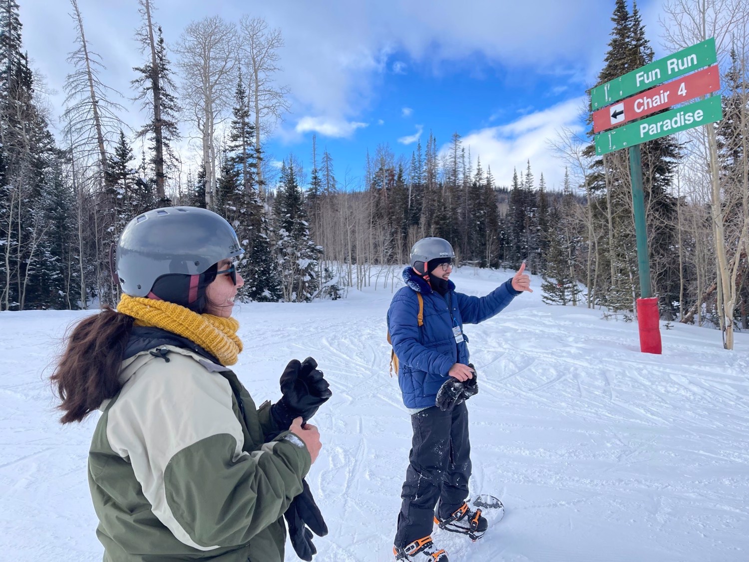Two helmeted club members pause beside a trail sign on a blue-sky snow day.