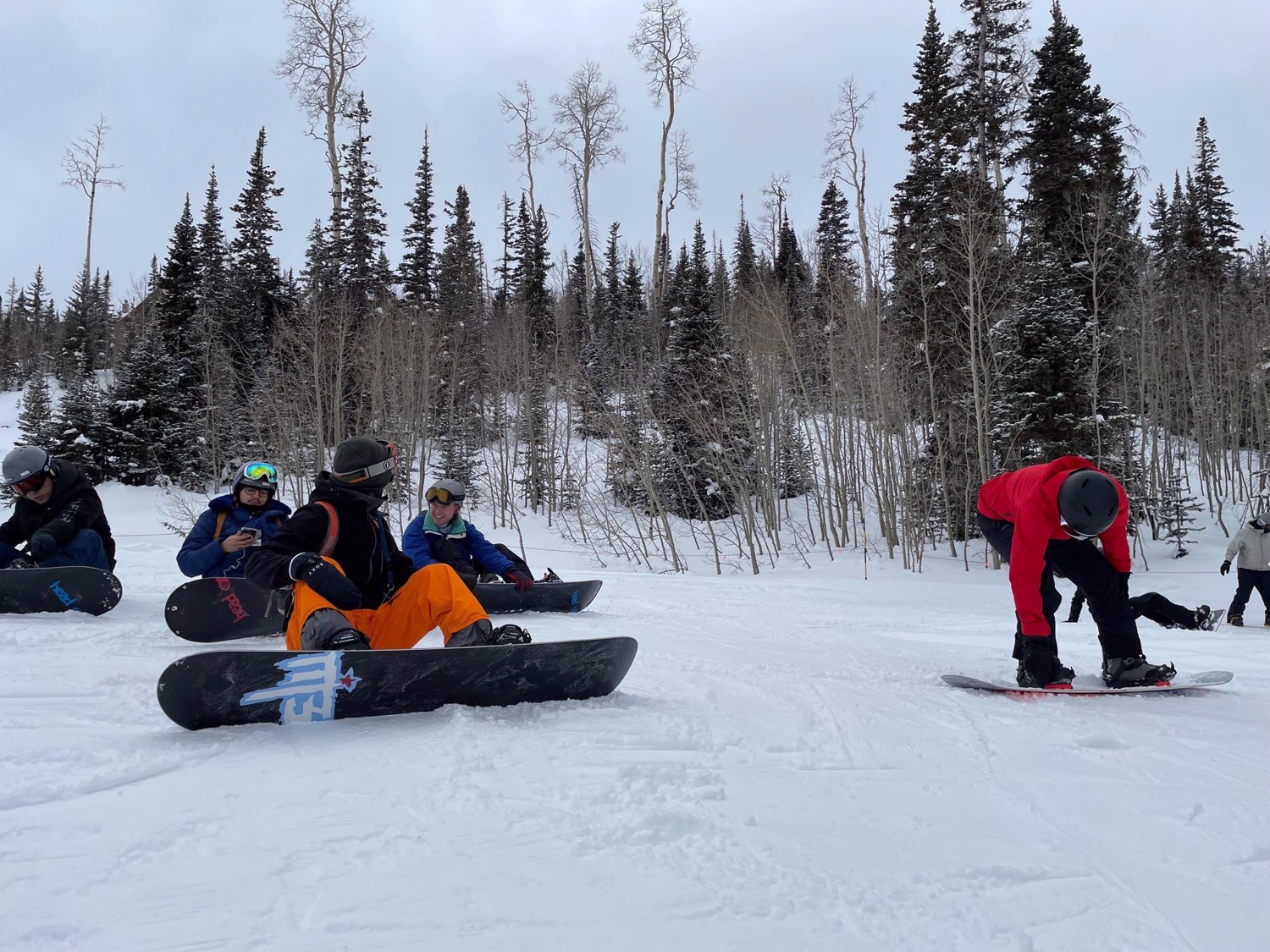 Club members rest with snowboards on a snowy trail surrounded by trees.