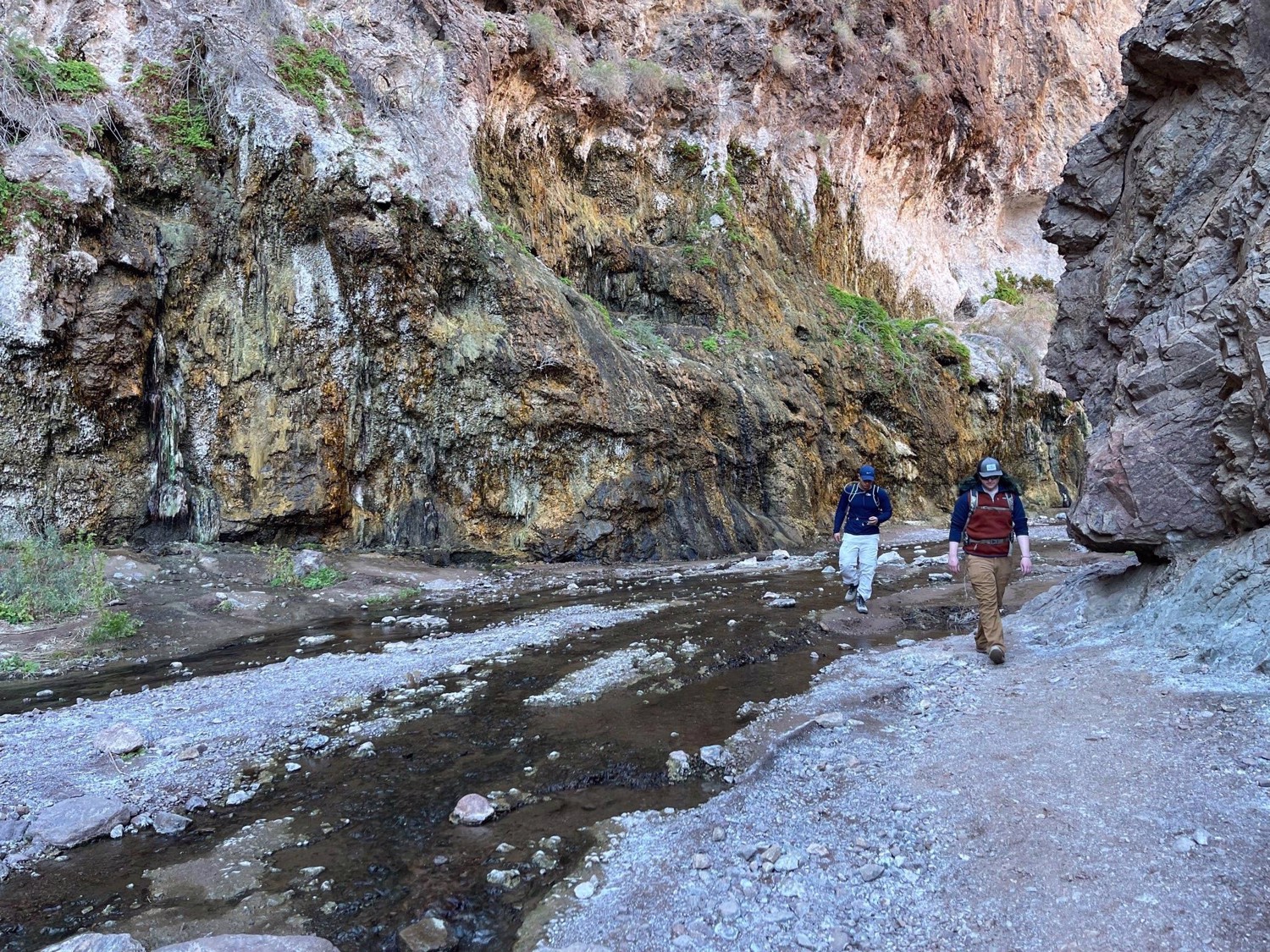 Two hikers walk beside an icy stream at the bottom of a rocky canyon.