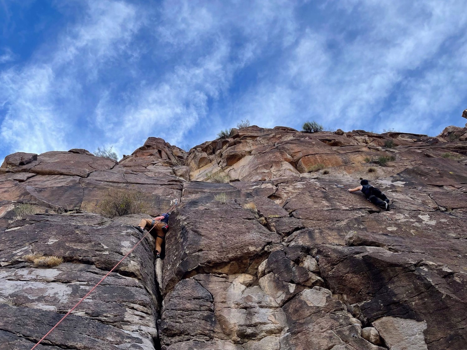 Two climbers move across a broad rock face beneath a blue, cloud-filled sky.